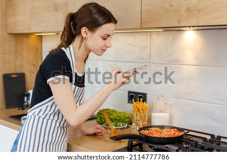 Young woman blogger in apron taking photo with phone while cooking food at kitchen. Preparing meal with frying pan on gas stove. Concept of domestic lifestyle, housewife leisure and culinary blogging.