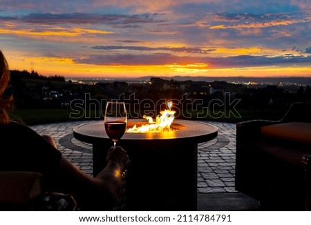 A woman drinks a glass of wine in front of a fire pit on the luxury patio of a hillside home overlooking city lights.	
