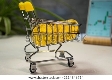 A shopping cart with Bitcoin coins on the background of a flower and a smartphone with a graph of the BTC cryptocurrency exchange rate. Blockchain technology. Bitcoin in everyday life