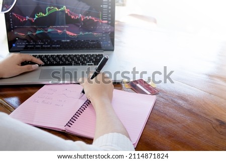 Using a computer to view stock market graphs and write them down in a paper notebook makes financial plans on a wooden table.
