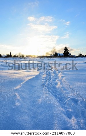 Winter landscape on frosty sunny morning. Snow-covered path with fresh footprints leads to houses in background. Sun's rays glide over snow. Vertical photo