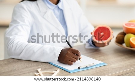 African american female nutritionist holding grapefruit, making meal plan for client at weight loss clinic, writing prescription for patient at office, closeup, crop