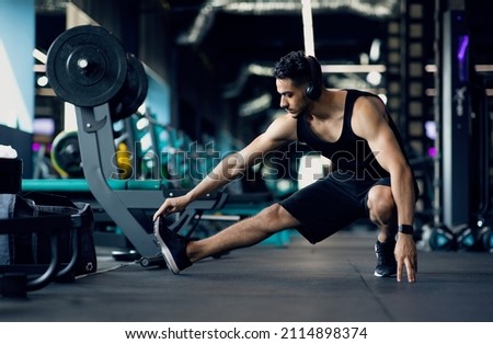 Handsome Young Middle Eastern Male Athlete Warming Up Before Training At Gym, Motivated Millennial Arab Man Stretching Leg Muscles, Preparing For Fitness Workout In modern Sport Club, Copy Space Royalty-Free Stock Photo #2114898374