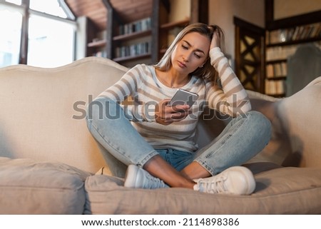 Worried lady looking at smartphone screen, sitting on couch at home, concerned middle aged woman waiting for important call, reading unpleasant sms, spending time holding phone in living room Royalty-Free Stock Photo #2114898386