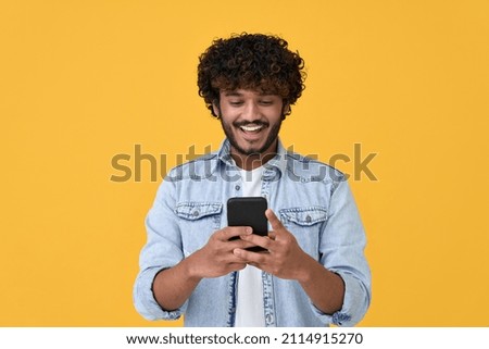 Happy indian young man using cell phone isolated on yellow background. Smiling ethnic hipster guy holding smartphone playing game in app, dating online, buying online in e commerce store on cellphone.