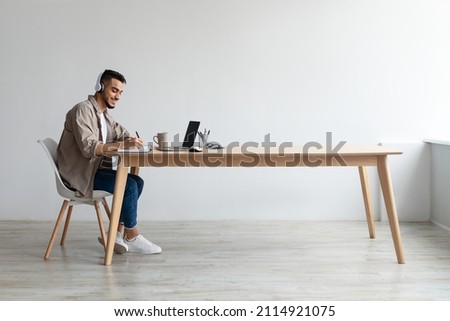 Profile portrait of young smiling Arab man in wireless headset using pc sitting at desk, writing in notebook. Cheerful guy browsing internet, watching webinar studying online, free copy space