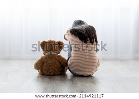 Back view of sad long-haired little girl in casual outfit sitting with toy teddy bear and crying, feeling down, upset kid got punished after quarrel with parents, copy space, home interior Royalty-Free Stock Photo #2114921117