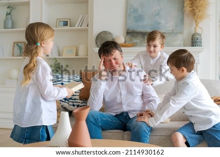 Tired father, sitting on couch in living room, feels annoyed and exhausted while noisy daughters and sons run screaming and demanding attention from dad. Overly active hyperactive children need rest Royalty-Free Stock Photo #2114930162
