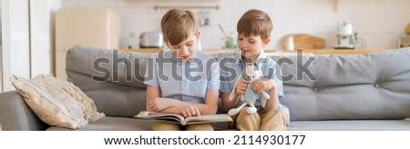 Teach your child to read book as teenager. Caucasian child looking into book while studying with teenager on couch in living room at home. Elder brother is reading book in living room on sofa brother
