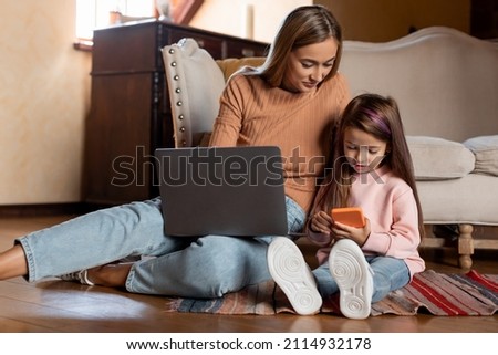 Happy adult lady sitting on floor using laptop relaxing with preschooler daughter holding smartphone have fun together, smiling woman and little girl enjoy weekend at home busy with gadgets