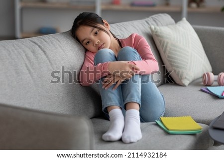 Upset asian schooler chinese girl in casual sitting on couch with notepads, hugging her knees, doing homework, having difficulties with education, got bored, home interior, copy space Royalty-Free Stock Photo #2114932184