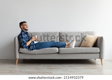 Rest Concept. Happy Arab guy drinking coffee sitting on comfortable couch at home in living room. Cheerful casual man relaxing on sofa, enjoying weekend free time or break from work, full body length Royalty-Free Stock Photo #2114932379