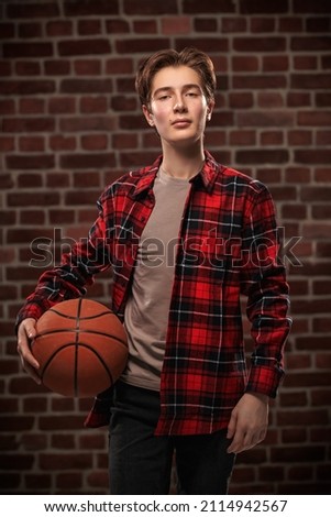 A modern teenager boy in casual clothes stands with a basketball against a brick wall. Sports, hobbies. Active lifestyle.