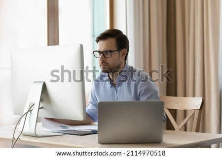 Concentrated young businessman in eyeglasses working on different devices, analyzing information, considering problem solution, creating new online project or doing economic research at home office.