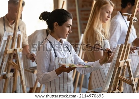 Focused inspired Indian artistic class student girl drawing in paints on canvas, taking lesson with group, holding, using artist tools, working on artwork in studio. Creative hobby concept