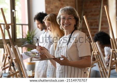 Happy senior retired student of art school drawing in class, using artist tools, canvas, standing at easel, looking at camera, smiling, laughing. Head shot portrait in artistic studio Royalty-Free Stock Photo #2114970707