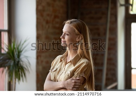 Peaceful thoughtful long haired young woman in casual looking at window away with folded hands, standing in loft space home, thinking, dreaming, smiling at good thoughts, pondering on future plans