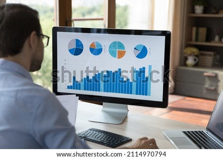 Rear view concentrated millennial businessman in glasses looking computer monitor, analyzing graphs and charts with online sales, project statistics data, developing marketing strategy at home office. Royalty-Free Stock Photo #2114970794