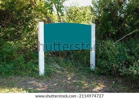 Blank green wooden sign on white posts with large bushes in the background