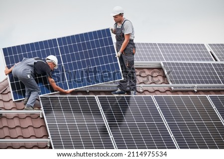 Men technicians carrying photovoltaic solar moduls on roof of house. Engineers in helmets installing solar panel system outdoors. Concept of alternative and renewable energy. Royalty-Free Stock Photo #2114975534