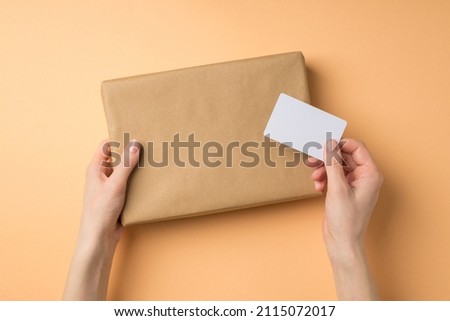 First person top view photo of young woman's hands holding craft paper giftbox and business card on isolated beige background with copyspace
