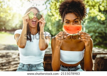 joiful multicultural young adult girlfriends sitting countryside under the trees eating fresh watermelon and hiding the faces with slices and peel - genz women sustainable lifestyle Royalty-Free Stock Photo #2115078983