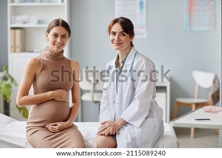 Portrait of pregnant woman and her gynecologist smiling at camera together while sitting on couch at doctors office