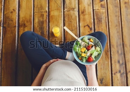 View from above of pregnant woman with fresh salad bowl and orange juice, expectant mother having healthy lunch while sitting on wooden floor in lotus pose, cropped. Nutrition during pregnancy Royalty-Free Stock Photo #2115103016