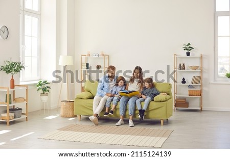 Happy family spending time at home. Mum, dad and little kids reading book while sitting together on green sofa in room with light walls, beige rug and simple brown shelves in their big new house
