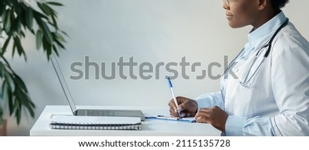African american female doctor making note in checklist clipboard looking at laptop screen sitting at desk works on document prescription or check medical appointment list. Closeup side view shot Royalty-Free Stock Photo #2115135728