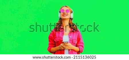 Portrait of stylish happy smiling young woman in headphones with smartphone listening to music wearing pink jacket, sunglasses on green background
