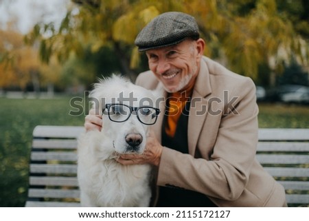 Happy senior man looking at camera and embracing his dog wearing glasses on bench outdoors in city.