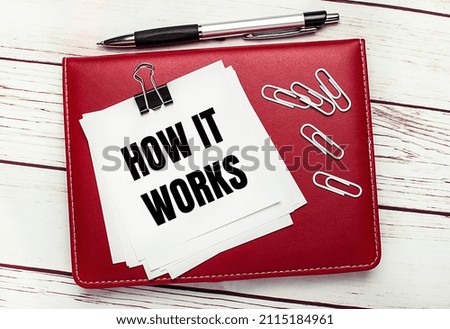 On a light wooden background, a burgundy pen and notebook. On the notebook has white paper clips and white paper with the text HOW IT WORKS. Business concept