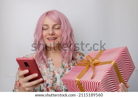 Pink hair girl holding surprise gift box with gold bow buys on the internet uses smartphone. Consumerism, sale, purchases, shopping, lifestyle concept.