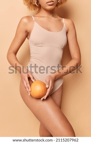 Vertical shot of faceless healthy woman in brown underwear holds orange over body has clean healthy skin without cellulite slender legs demonstrates perfect figure after liposuction poses indoor