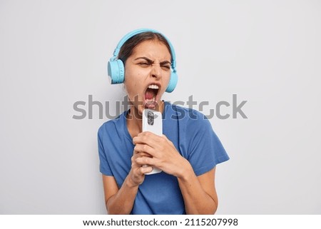 Carefree dark haired female singer keeps mouth widely opened listens music via headphones holds smartphone as if microphone enjoys loud sound dressed casually isolated over white background.