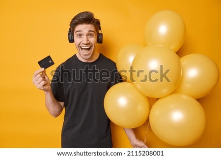 Joyful man happy to get big sum of money on his bank account ready to celebrate birthday holds bunch of inflated balloons and credit card listens music via headphones isolated over yellow wall