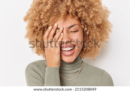 Overjoyed curly haired woman makes face palm laughs happily expresses positive emotions wears casual turtleneck isolatedover white background hears funny story from friend. Happiness concept
