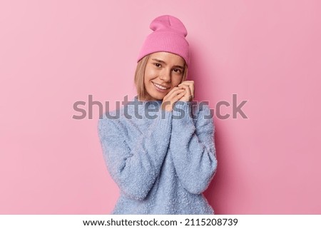 Waist up shot of pleased tender woman keeps hands near face smiles happily wears soft jumper and hat being in good mood isolated over pink background. People emotions face expressions concept