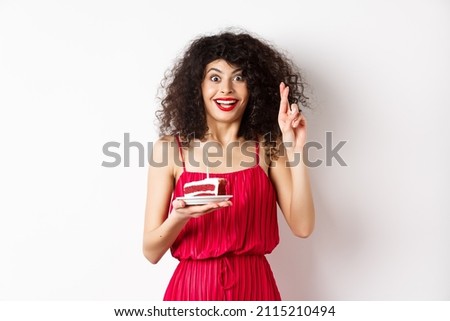 Excited birthday girl in red dress, cross fingers while making wish and blowing candle on bday cake, smiling happy, white background