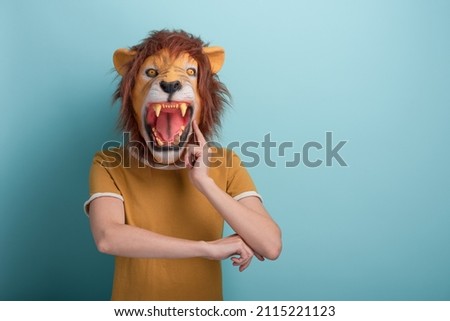 Young woman in lion mask with hand on chin, thinking, pensive expression, isolated on blue background with copy-space.