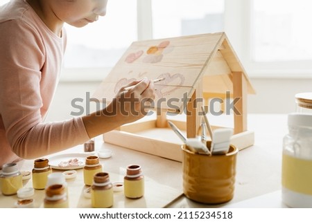 Caucasian girl kid colors wooden bird feeder, winter bird care and children's creativity at home. High quality photo Royalty-Free Stock Photo #2115234518