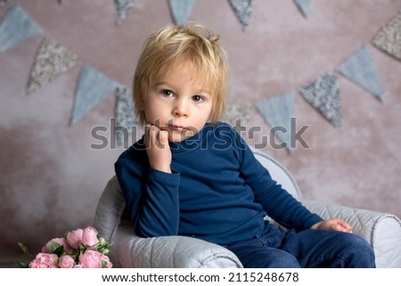 Cute blond toddler child, boy, sitting on little baby armchair, reading a book and holding flowers, studio indoors shot