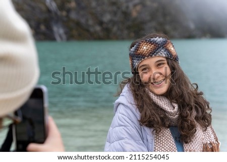 Smiling young woman posing while a friend taking a photo in front of Llanganuco lagoon in Peru