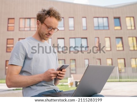 Young freelancer using laptop computer, holding mobile phone working online sitting at workplace. Student studying, distance learning sitting in university campus, education concept  