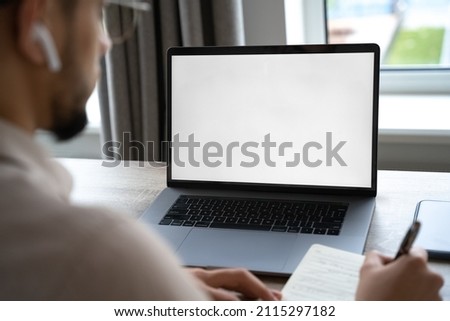 Young man working on laptop with blank copy space screen from home office. Over shoulder view on student or businessman writing notes in notebook looking at portable computer monitor