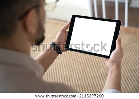 Closeup of Indian man holding digital tablet working online sitting on sofa at home office. Over shoulder view of male businessman freelancer or student watching training tutorial, webinar Royalty-Free Stock Photo #2115297260