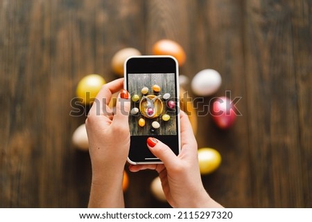 Woman holding mobile phone and making photo of eggs painted for Easter isolated on wooden background. Easter eggs flat lay on rustic table. Easter concepts