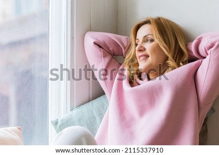 A mature woman in a pink hoodie sits on the windowsill with her arms folded behind her head and enjoys the moment. Slow life concept and digital detox Royalty-Free Stock Photo #2115337910