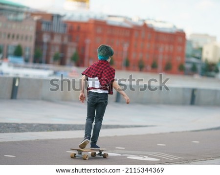 Skateboarder riding on the road in the Moscow public park. The ends of his hair are dyed blue. He is training. Active lifestyle and youth fashion concept. Back, rear view